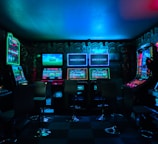 Dimly lit gaming arcade featuring a variety of brightly illuminated slot machines and video gaming screens. Neon signs and colorful displays create a vibrant atmosphere. The arrangement of gaming machines suggests an inviting space for entertainment, with chairs positioned for players.